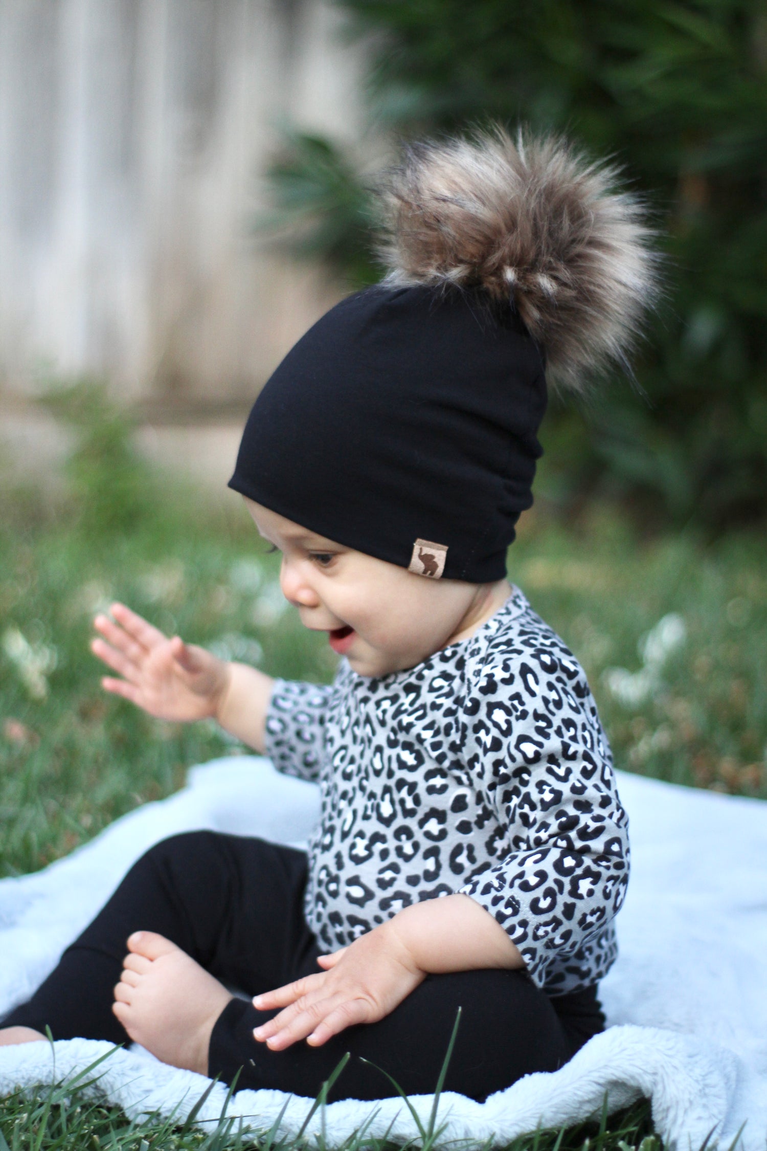 baby sitting on a blanket wearing a black pom pom beanie and leopard onsie