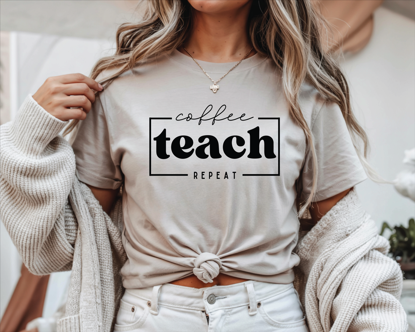 A woman wearing a light beige graphic t-shirt with the words 'coffee teach REPEAT' printed in black. She has a chunky knit cream cardigan draped off her shoulders and is wearing white jeans.