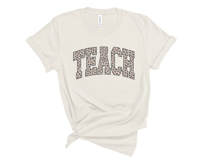 An off-white short-sleeve t-shirt with the word 'TEACH' printed across the chest in a leopard print pattern. The t-shirt is styled with a knot at the bottom hem and is photographed flat against a solid white background.